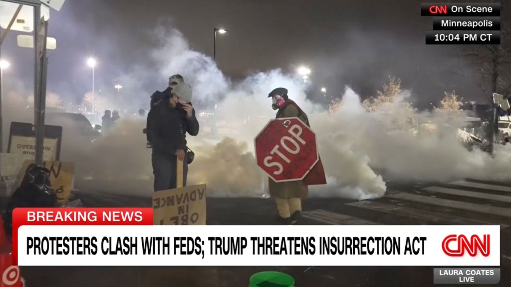 People covered in smoke, one holding a stop sign, at a protest where federal agents deployed tear gas.