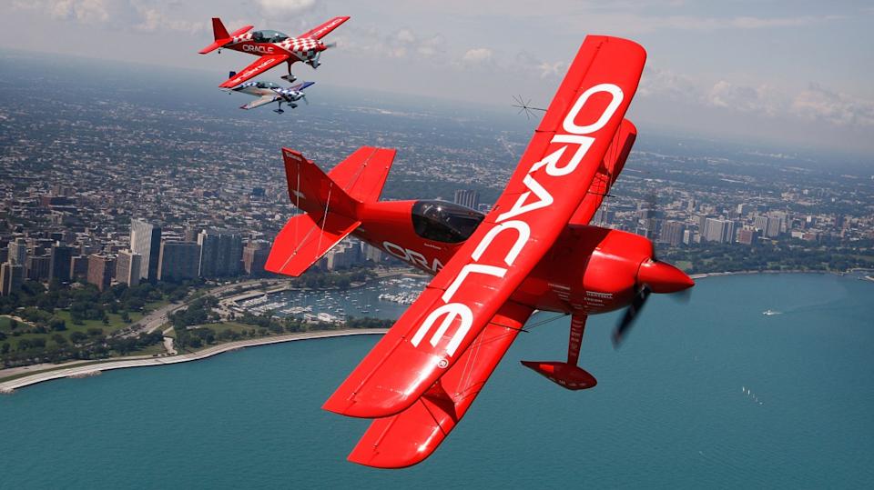 Three red biplanes, two smaller ones in the distance and a large one prominently in the foreground, with 'ORACLE' written in white letters on their wings. The planes are flying over a sprawling city skyline beside a large body of blue water with boats. The sky is partly cloudy.