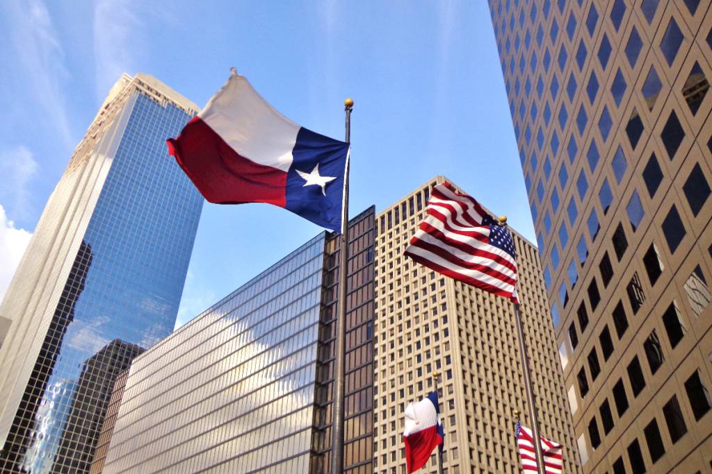 Image shows Texas and U.S. flags flying with buildings in the background.