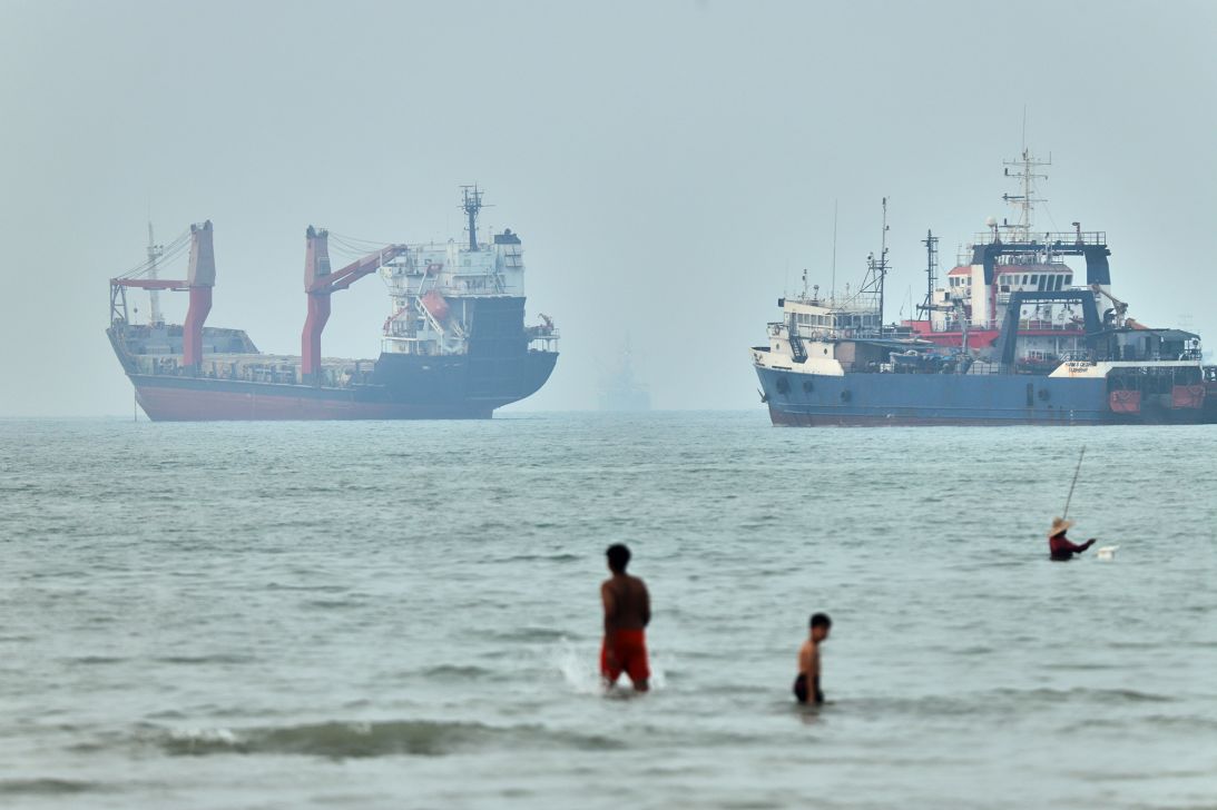 Children wade in the water with cargo ships at anchor in the background and a fisherman nearby, in the Strait of Hormuz off Bandar Abbas, Iran, Tuesday, June 30, 2026.