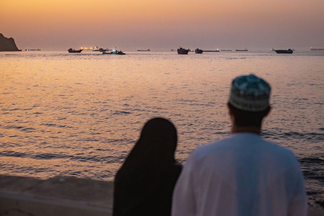 A couple watch oil tankers, general cargo ships, bulk carriers, and fishing boats anchored in the waters off Muscat near the Strait of Hormuz on June 18, 2026 in Muscat, Oman.